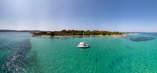 Catamaran anchored in crystal clear turquoise water near a forested Greek island beach, surrounded by small boats on a sunny summer day. Aerial view