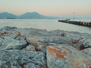 Peaceful coastal landscape with large rocks in the foreground, calm sea, distant mountains and an empty wooden pier stretching into the water. Bright natural daylight. Soft focus