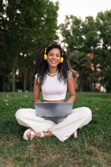 Obraz premium Smiling young woman working with a laptop and headphones in a park surrounded by many trees