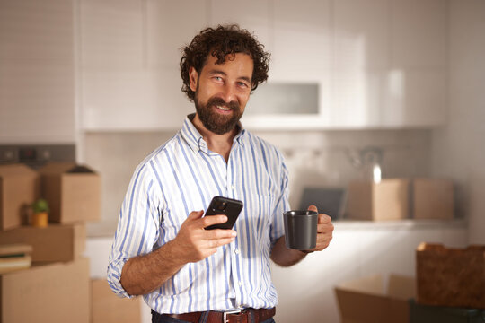 A man stands in a bright kitchen filled with moving boxes, holding a coffee cup in one hand and a smartphone in the other, preparing for the move to his new home.