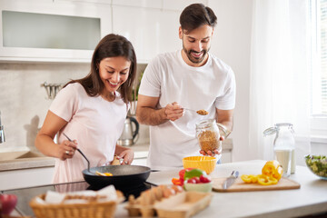 In a modern kitchen filled with natural light, a couple happily prepares a healthy meal. The woman stirs a pan while the man adds ingredients from a jar, both smiling and engaged.