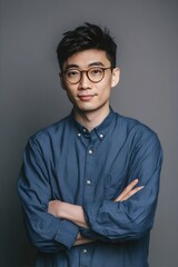 Fototapeta premium A man in glasses, wearing a blue shirt, stands confidently with arms crossed against a grey background.