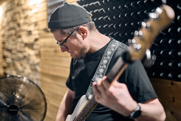 Young male guitarist with dark hair and glasses plays bass guitar in acoustically treated studio. Soft lighting enhances concentration. Wooden and foam backgrounds create warm, creative ambiance