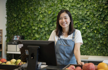 Asian female fruit store owner smiles confidently behind counter wearing denim apron. Modern retail setup with green wall background creates natural indoor ambiance. Focus on fresh produce, healthy