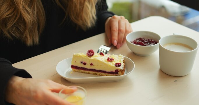 WARSAW, POLAND - MARCH 10 2023: Woman pours sauce onto cheesecake with berries sitting at cafe table. Lady adds creamy topping to dessert for extra tenderness before eating with coffee