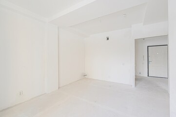 bright, empty room with white walls, ceiling, and unfinished floor. A doorway leads to a hall with an entrance door. The space is minimal and appears to be under construction