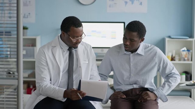 Medium shot of Black male doctor handing patient clipboard with official medical document or consent form for signing during healthcare appointment, indicating acknowledgment of medical information