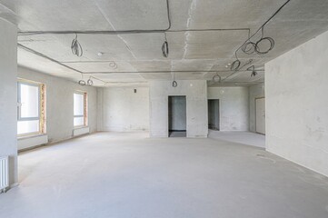spacious, empty room under construction. Cement walls and floor, exposed wiring on the ceiling, and window openings await finishing