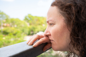 Side view portrait of a sad single woman looking down from a balcony of a house with an urban background