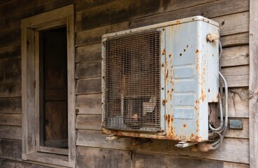 Rusty old air conditioner unit mounted on weathered wooden wall next to open doorway. Appliance shows signs of rust and wear, suggesting outdated technology and need for replacement or upgrade.