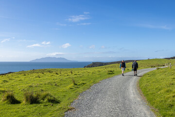 Two hikers walk along a gravel path on a sunny day in Tawharanui, New Zealand. They are enjoying the scenic views of the green hills and the ocean with an island in the distance.