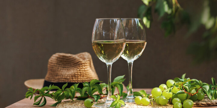Two glasses of chilled white wine with grapes and straw hat on sunlit outdoor setting..