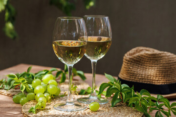 Two white wine glasses with green grapes and hat on wooden table with vines and soft light..