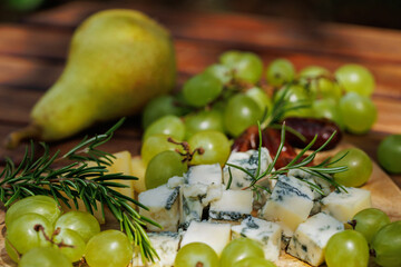 Rustic wooden platter with cheese, green grapes, dates, rosemary and ripe pear on terrace table..