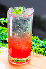 Refreshing red cocktail with ice and mint leaves served in a clear glass on a wooden surface with green foliage background.
