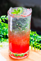 Refreshing red cocktail with ice and mint leaves served in a clear glass on a wooden surface with green foliage background.