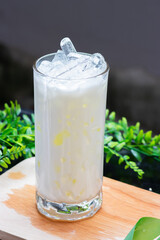 A cold glass of coconut milk with sweet corn and ice cubes, served on a wooden surface with a natural green background.