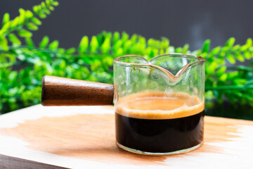 Freshly brewed espresso in a clear glass cup with a wooden handle, showcasing rich crema, placed on a wooden board with green foliage in the background.