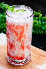 A vibrant iced strawberry milk drink swirled with red syrup, topped with fresh mint and ice, served in a clear glass on a wooden surface with greenery.