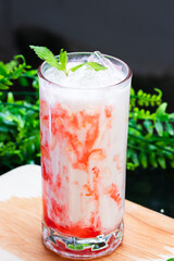 A vibrant iced strawberry milk drink swirled with red syrup, topped with fresh mint and ice, served in a clear glass on a wooden surface with greenery.