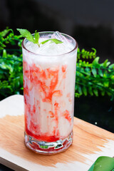 A vibrant iced strawberry milk drink swirled with red syrup, topped with fresh mint and ice, served in a clear glass on a wooden surface with greenery.