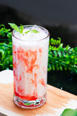 A vibrant iced strawberry milk drink swirled with red syrup, topped with fresh mint and ice, served in a clear glass on a wooden surface with greenery.