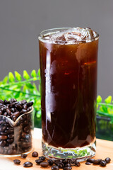 A refreshing iced black coffee in a tall glass with ice cubes, served alongside a clear container filled with roasted coffee beans on a wooden board.