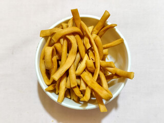 The white bowl contains the Indonesian cash food ladrang snack, which has a savory golden colored taste in the shape of a stick or pipe, served on a light colored cloth.