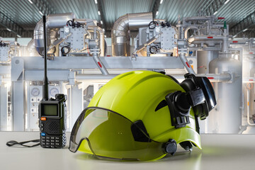Protective helmet, goggles, and radio on table with industrial equipment in background, representing safety in engineering, maintenance, and factory operations. Engineering, Industry, Safety.