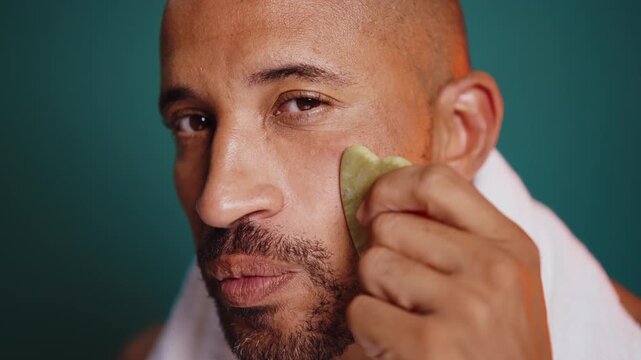 Attractive bald man with a white towel around his neck smiles while applying under-eye patches, showcasing a skincare routine against a brown backdrop