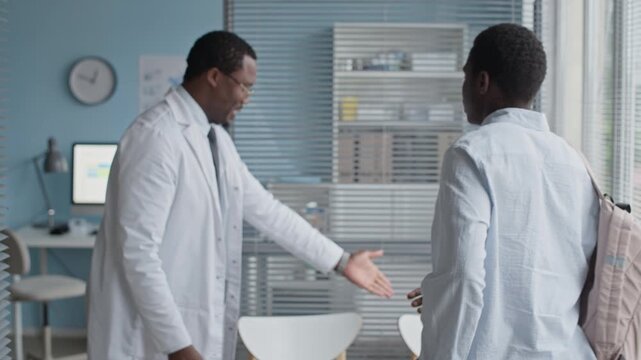 Medium shot of confident Black male physician greeting patient with handshake at office entrance, offering courteous and professional start to medical consultation