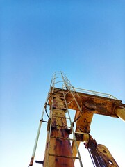 A rusted yellow industrial ladder and crane structure stand against a clear blue sky. The metal shows significant signs of age and corrosion. The perspective is low, looking up.
