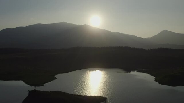 Reverse drone flyover above the island in Llyn y Dywarchen lake at sunrise with Snowdon mountain in the distance. Scenic North Wales landscape linked to Welsh folklore and Tylwyth Teg legends.