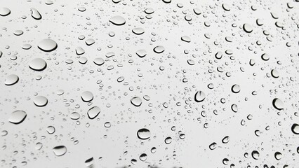 Water droplets on glass vary in size and shape, adhering due to surface tension. These droplets create a natural pattern against a blurred, light gray background.