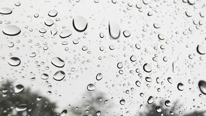 Water droplets on glass vary in size and shape, adhering due to surface tension. These droplets create a natural pattern against a blurred, light gray background.