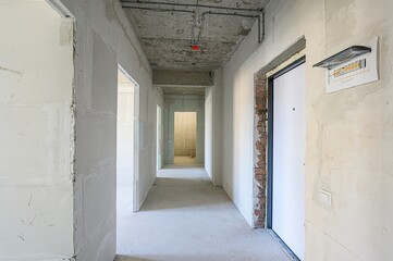 unfinished hallway with bare concrete floors and walls, exposed wiring, and doorways leading to other rooms. A white door stands out on the right with an electrical panel nearby