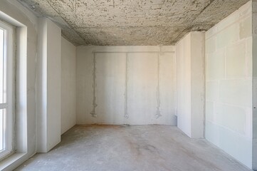 Empty room under construction. Concrete ceiling, unfinished walls, and visible electrical wiring create a raw, industrial look. A window provides natural light
