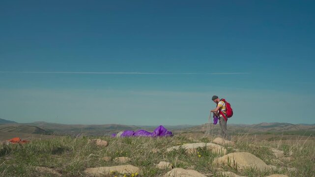A paraglider man with a parachute preparing equipment on a hilltop for flight in wilderness area. Cross country pilot enjoying extreme sports.