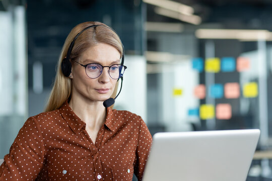 A female customer service representative wears a headset while working on her laptop in a modern office setting.