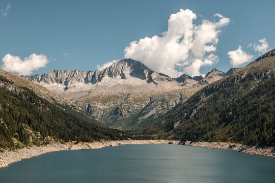 A scenic view of a blue lake surrounded by forested mountains under a partly cloudy sky. Val di Fumo, Chiese river, Carr&egrave; Alto, Trentino. The water level is low, exposing a rocky shoreline.