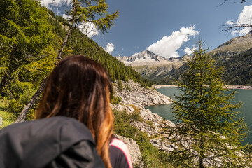 A woman with a backpack looks out over a lake and mountains. Val di Fumo, Chiese river, Carrè Alto, Trentino. The scene evokes a sense of adventure and appreciation for the natural world.