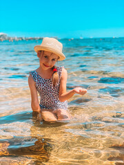 A young girl in a hat sits in shallow water on a sandy beach, surrounded by clear turquoise waves. Peaceful summer scene with bright sunlight, relaxing atmosphere, and tropical seaside vibes.
