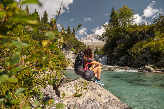 A woman with a backpack sits on a rock by a clear turquoise river, looking at a waterfall and mountain. Val di Fumo, Chiese river, Carr&egrave; Alto, Trentino. She is enjoying the scenic view during a hike.