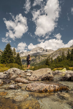 A hiker with a backpack stands on rocks in a stream, looking towards mountains under a cloudy sky. Val di Fumo, Chiese river, Carr&egrave; Alto, Trentino. The clear water flows around the rocks.