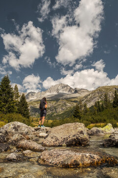 A woman with a backpack stands on rocks by a river, looking towards mountains. Val di Fumo, Chiese river, Carr&egrave; Alto, Trentino. 