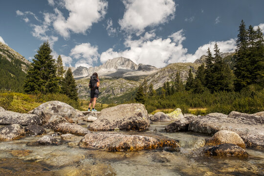 A woman with a backpack stands on rocks by a river, looking towards mountains. Val di Fumo, Chiese river, Carr&egrave; Alto, Trentino. 
