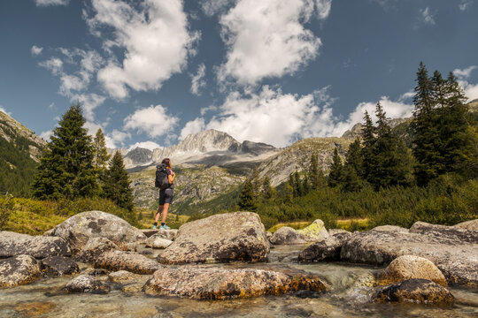 A woman with a backpack stands on rocks by a river, looking towards mountains. Val di Fumo, Chiese river, Carr&egrave; Alto, Trentino. 