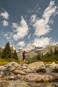 A woman with a backpack stands on rocks by a river, looking towards mountains. Val di Fumo, Chiese river, Carr&egrave; Alto, Trentino. 
