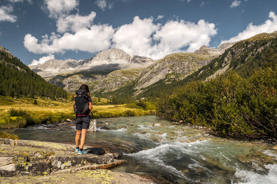 A woman with a backpack stands on rocks by a river, looking towards mountains. Val di Fumo, Chiese river, Carr&egrave; Alto, Trentino. 