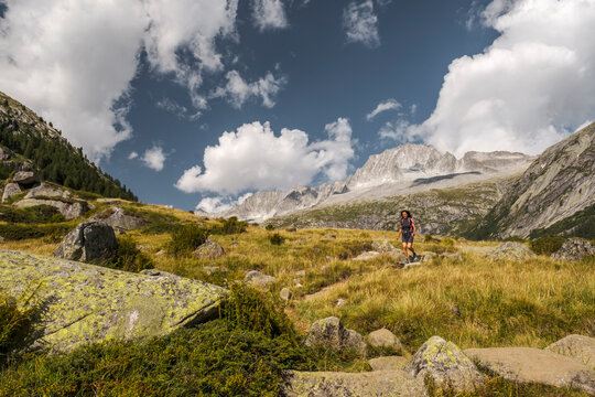 A woman hikes in a mountain landscape with a blue sky and clouds. Val di Fumo, Chiese river, Carr&egrave; Alto, Trentino. She is wearing hiking clothes and carrying a backpack.